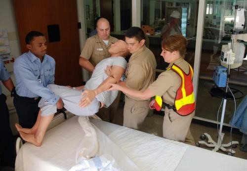 Four people in uniforms lift a medical training mannequin from a hospital bed in a clinical setting.