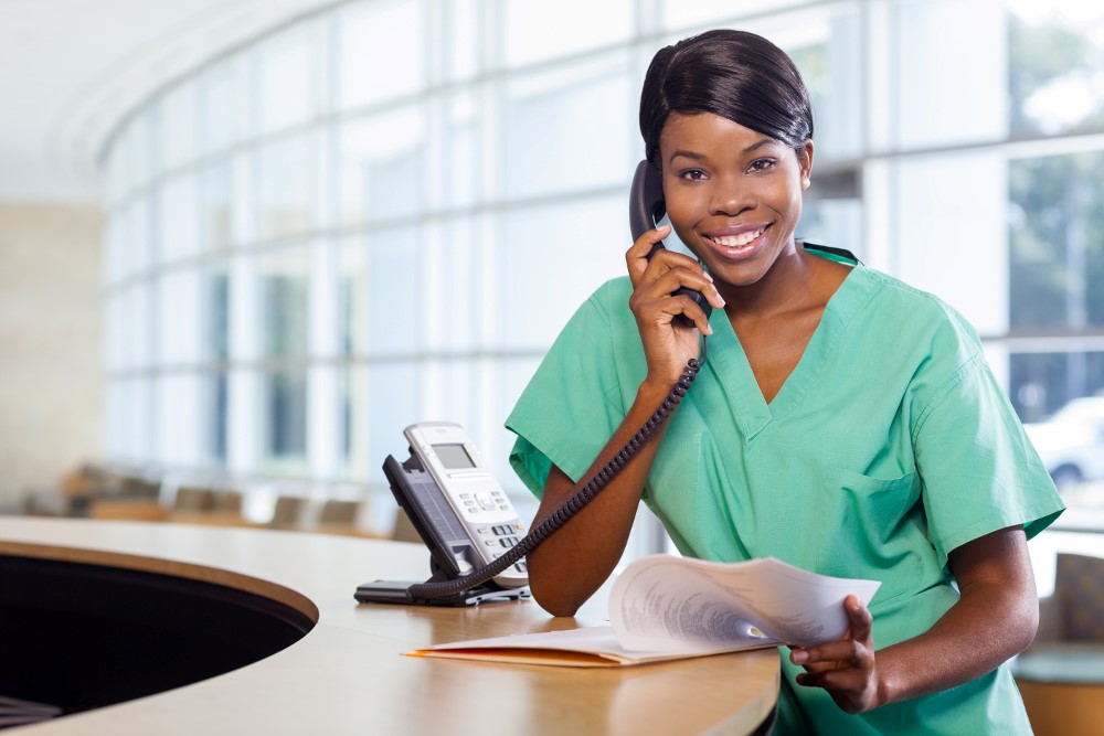 Nurse in green scrubs sitting at a desk, holding papers and talking on the phone in a bright office setting.