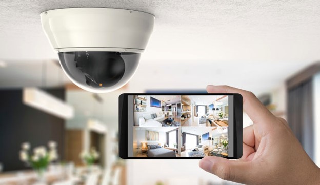 A hand holding a smartphone displays security camera footage; a dome security camera is mounted on the ceiling above.