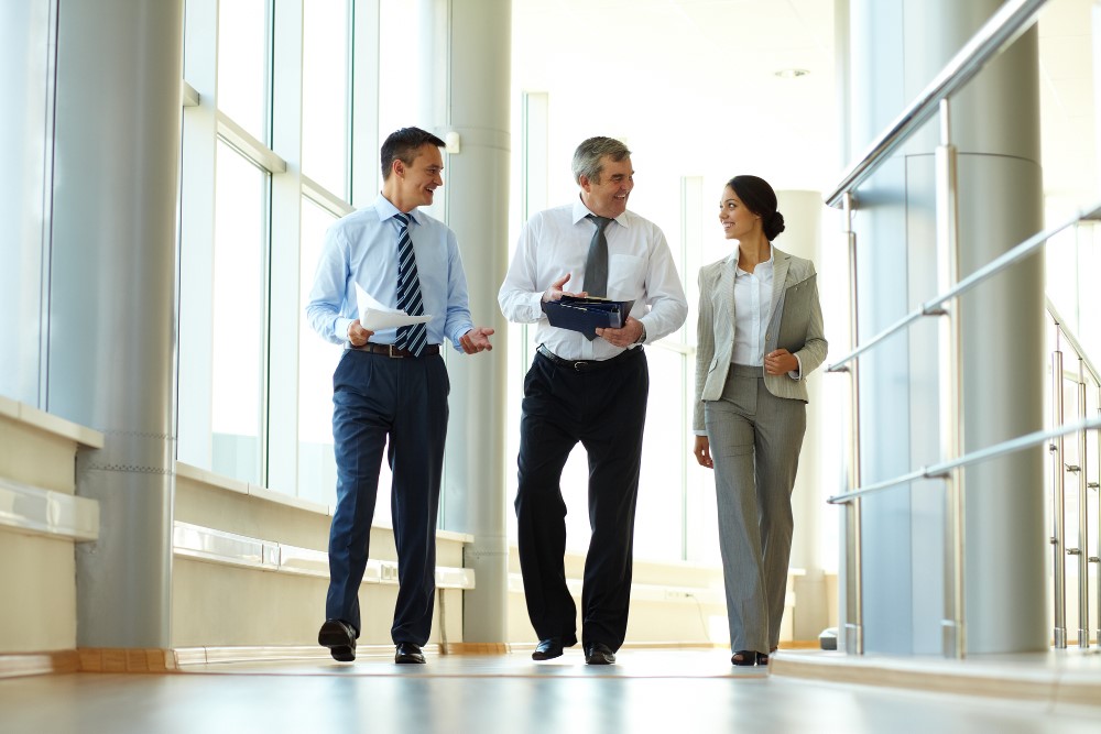 Three business professionals in formal attire walk and talk in a bright office hallway.